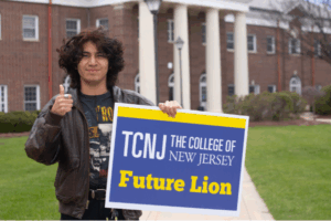 Student giving thumbs up and holding a blue sign that says TCNJ Future Lion
